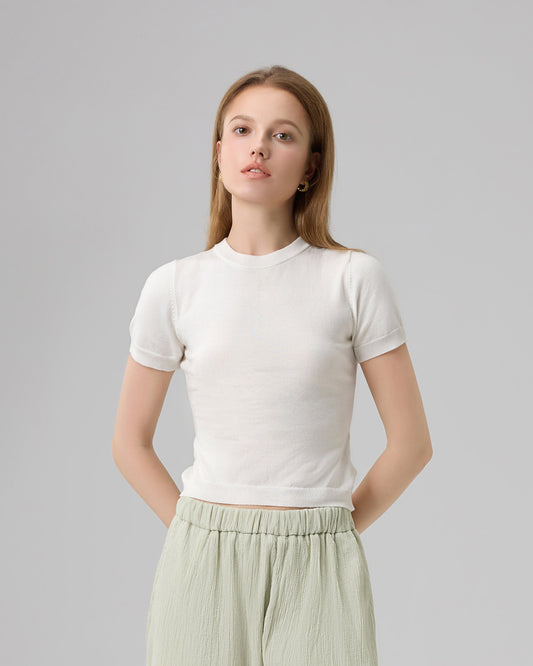 A woman modeling a white short-sleeve crop top and light sage green wide-leg pleated pants, paired with brown leather slide sandals, against a neutral light gray studio background.