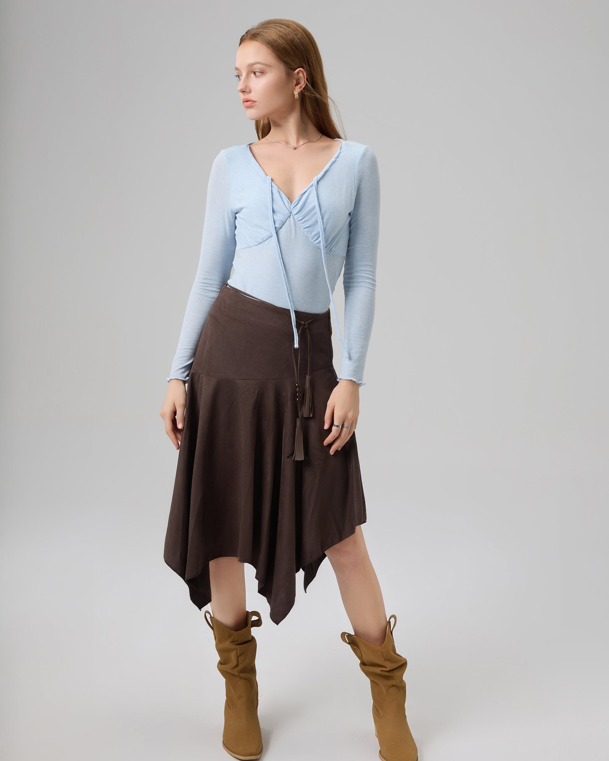 Woman wearing a light blue top, brown skirt, and brown boots on a gray background