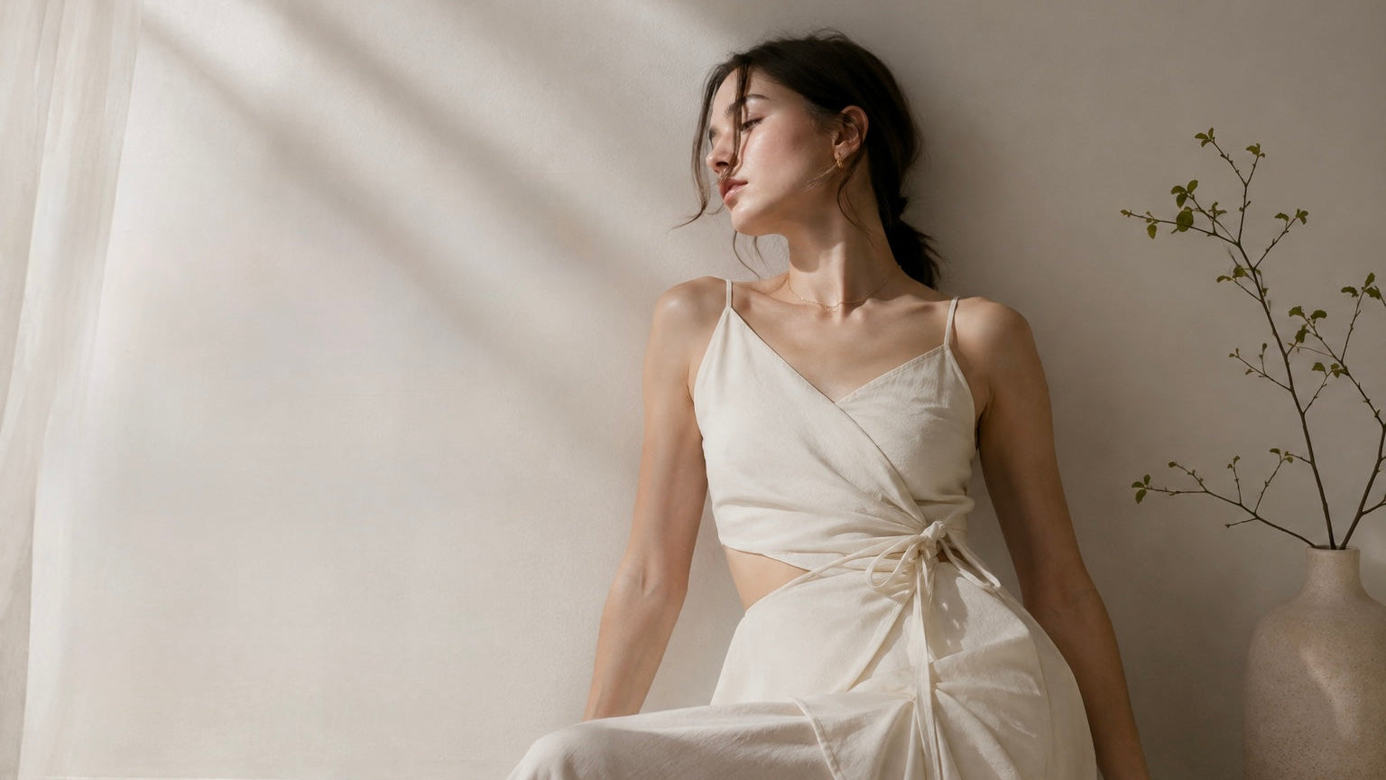 A woman wearing a cream-colored wrap-style midi dress with spaghetti straps and a side tie, sitting against a warm beige wall in a softly lit room with a vase of branches nearby.