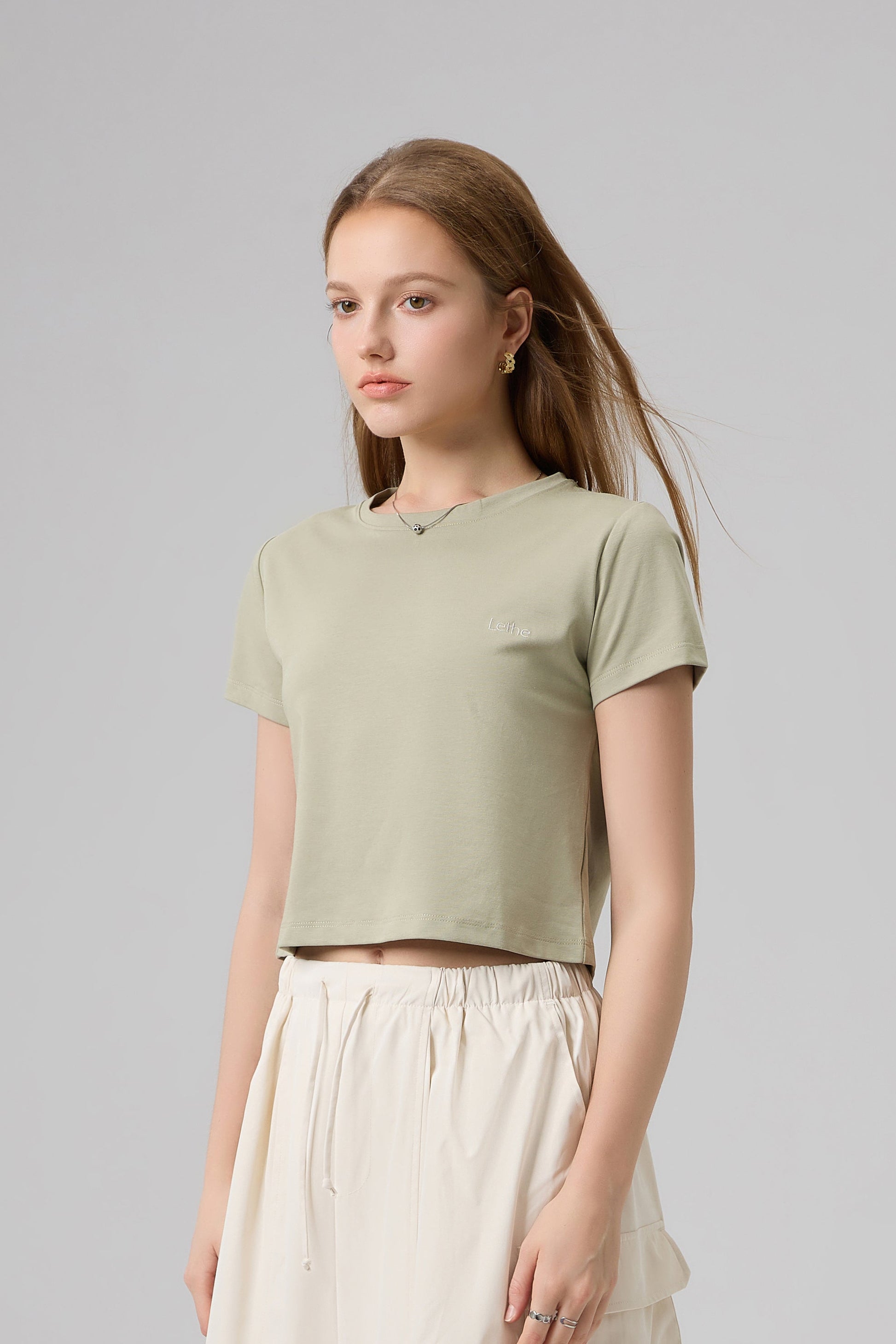 Model wearing The Lethe Logo Tee in Desert Taupe, short-sleeve cropped cotton t-shirt styled with cream drawstring skirt.