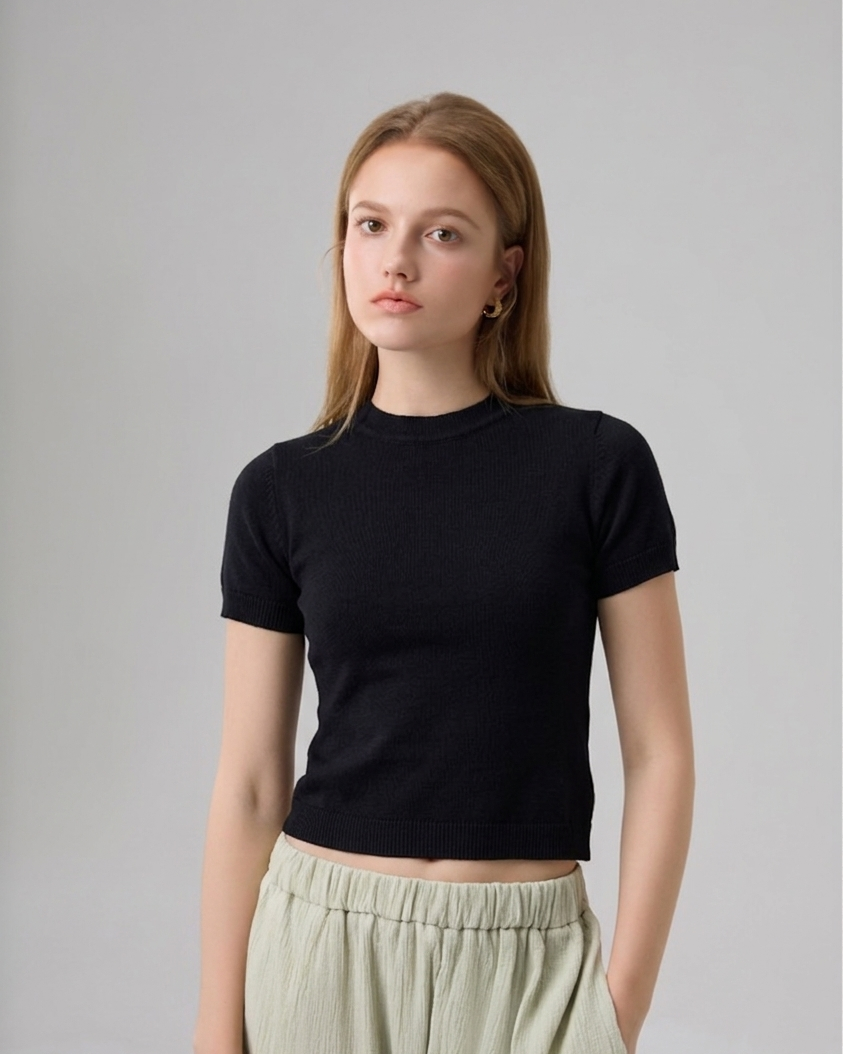 A woman modeling a black short-sleeve crewneck crop top paired with light sage green pleated trousers, photographed against a neutral light gray studio background.
