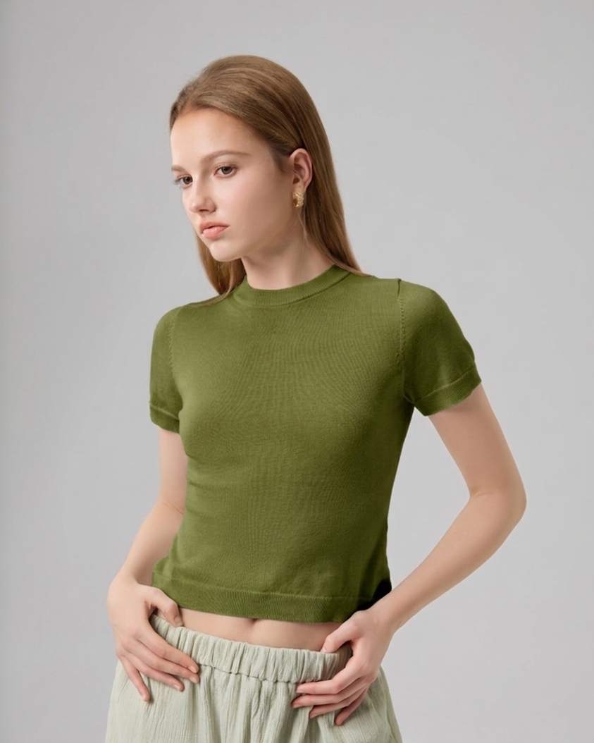 A woman modeling a fitted short-sleeve ribbed knit crop top in olive green, paired with light sage green pants, against a plain light gray studio background.