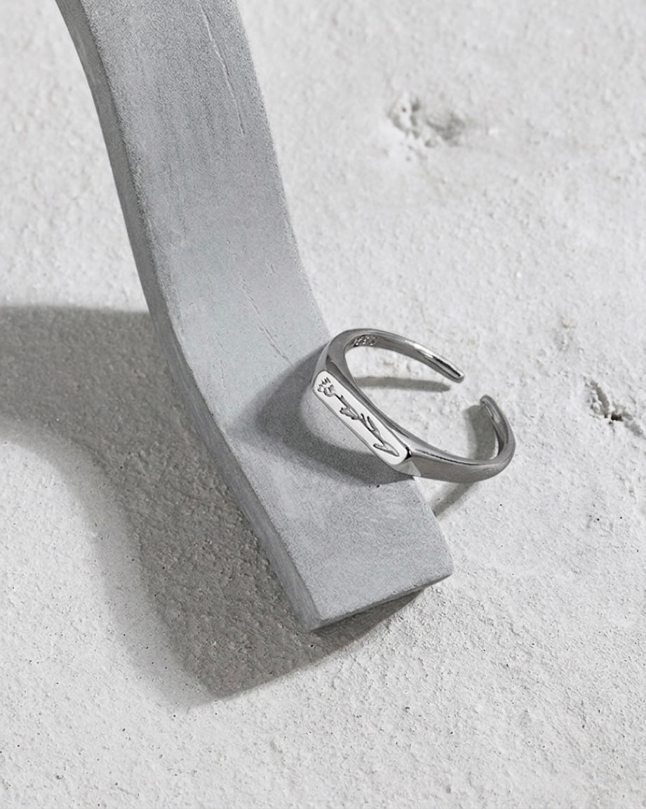 A silver open cuff ring with engraved script, displayed on a light gray velvet jewelry stand against a textured light gray background.