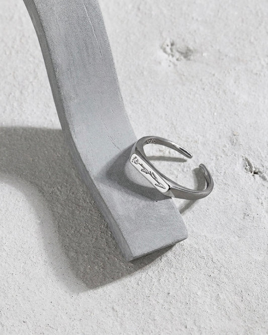 A silver open cuff ring with engraved script, displayed on a light gray velvet jewelry stand against a textured light gray background.