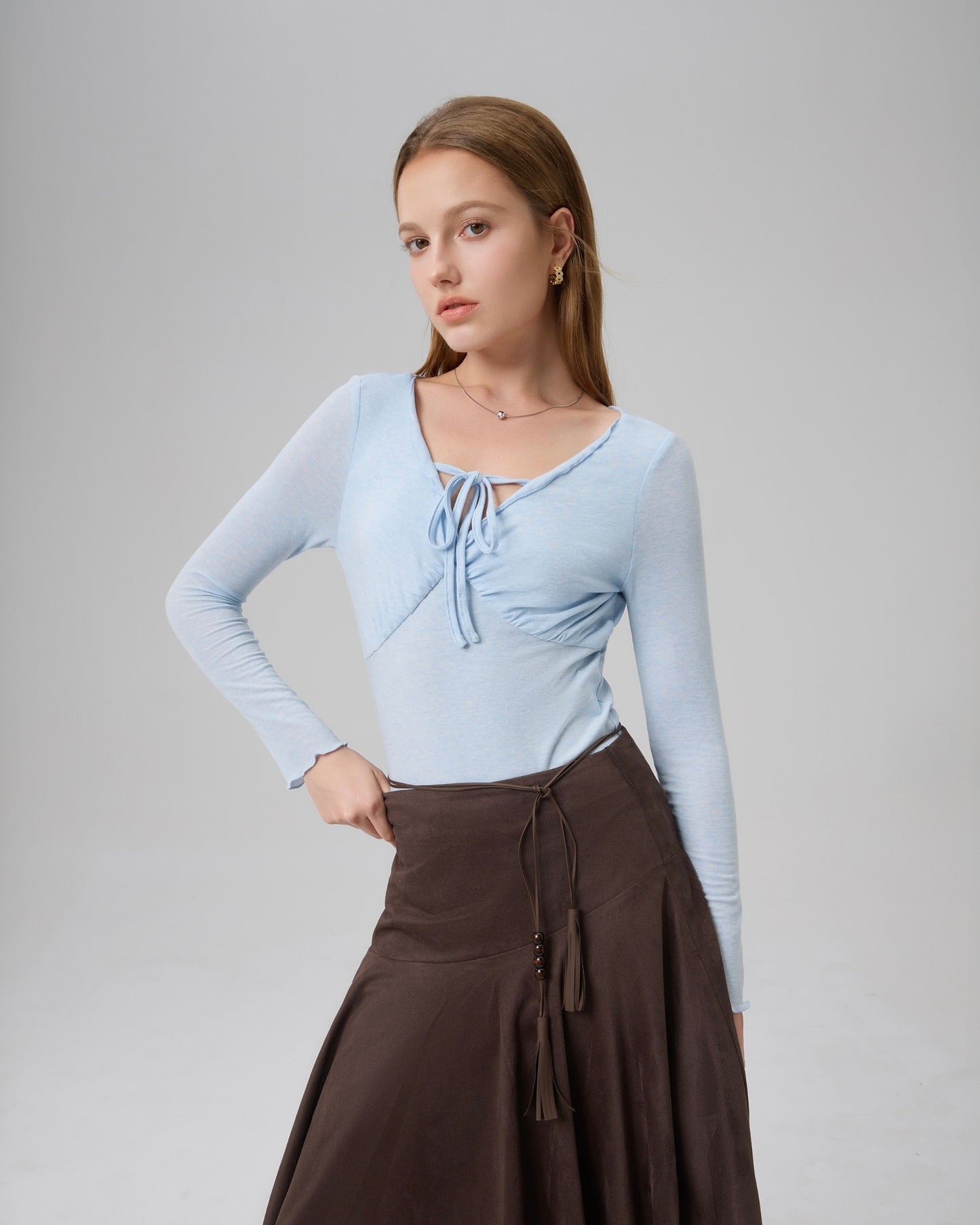 Model wearing The Knit Reverie Flowing Top in icy blue with a ruched front tie and V neckline, paired with a brown asymmetrical skirt.